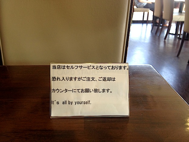Meaning, please seat yourself. Or suit yourself and sit alone. You're on your own.